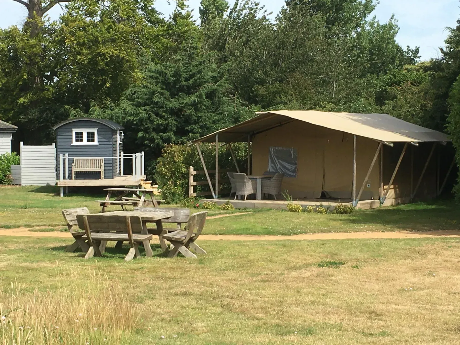 An open lawn with wooden picnic benches, a small shepherd’s hut and a canvas lodge set back under trees.