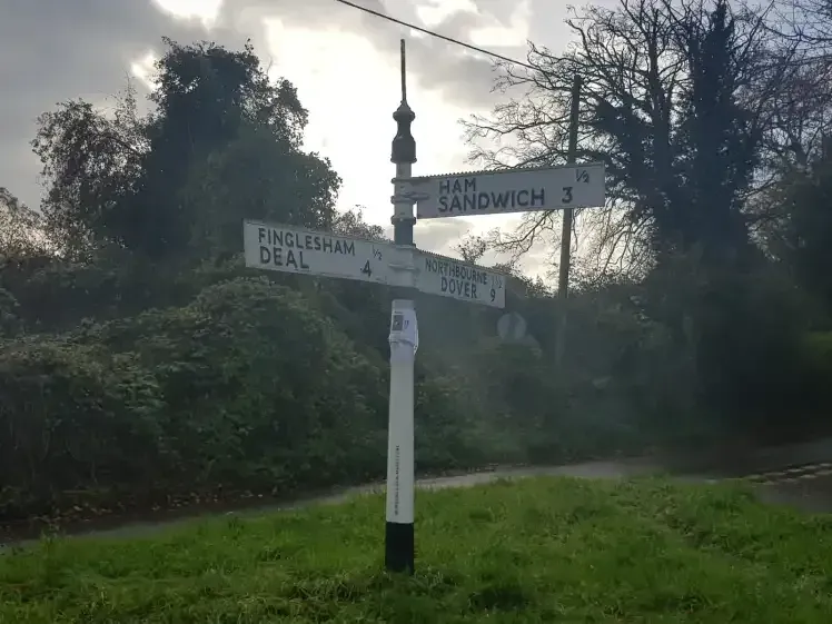 A white country fingerpost at a lane junction pointing to Ham, Sandwich, Finglesham, Deal and Dover.