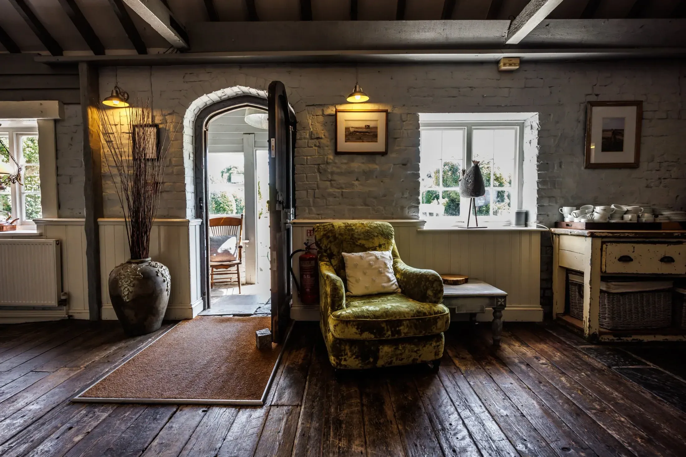 A sitting room with a bare board floor, a green velvet armchair beside a brick archway, and a plain kitchen dresser under a small window.